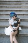 Desde arriba de mujer joven con el pelo azul corto y en vestido futurista de moda escuchando música con teléfono en las escaleras de la calle - foto de stock