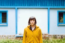 Jeune femme en manteau chaud jaune souriant et regardant la caméra tout en se tenant contre le bâtiment mural gris — Photo de stock