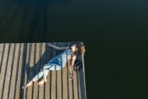 Top view of beautiful woman in black swimsuit and hat lying down on wooden pier of lake on clear blue sky and forest background — Stock Photo