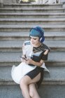 From above of young woman with short blue hair and in trendy futuristic dress listening to music with phone on street steps — Stock Photo