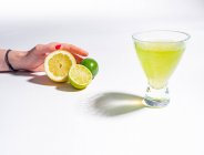 Mano femenina sosteniendo la mitad de limón y limas cerca del vaso de bebida de limonada amarilla sobre fondo blanco - foto de stock