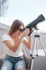 Young attractive woman sitting on chair and looking through telescope at sky — Stock Photo
