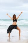 Attractive woman in black outfit dancing on sand near waving sea — Stock Photo