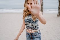 Cheerful blonde woman in colorful top and jean shorts blocking her face with hand on seashore — Stock Photo