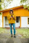 Mujer joven en traje casual sonriendo y navegando teléfono inteligente, mientras que de pie en el camino de baldosas fuera encantadora casa de campo en el día de otoño en el campo - foto de stock