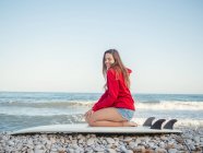 Seitenansicht der attraktiven lächelnden langhaarigen Frau in rotem Kapuzenpullover, die es genießt, auf einem Surfbrett am steinigen Meer zu sitzen — Stockfoto