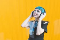 Joven alegre en vestido alternativo de moda sonriendo y escuchando música en auriculares contra la pared amarilla - foto de stock