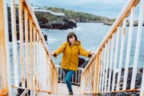 Молодая женщина в жёлтой куртке стоит на ржавой лестнице у залива с размахивающей морской водой — стоковое фото