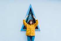 Junge Frau in gelbem warmen Mantel, lächelnd und in die Kamera blickend, während sie vor Dreiecksfenster und grauer Hauswand steht — Stockfoto