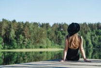 Обратный вид на неузнаваемую вдумчивую женщину в черном купальнике и шляпе, сидящую на деревянном пирсе и любующуюся видом на озеро на ясном голубом небе и лесном фоне — стоковое фото