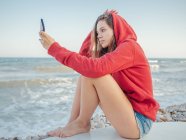 Молода усміхнена жінка з довгим волоссям у червоному светрі бере селфі на смартфон, лежачи на дошці для серфінгу на кам'яному узбережжі — стокове фото