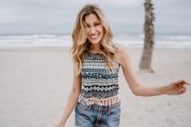 Fröhliche blonde Frau in buntem Top und Jeanshosen lächelnd und in die Kamera blickend, während sie sich am Strand entspannt — Stockfoto