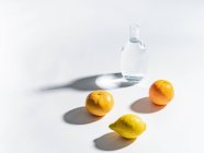 Arance mature e limone vicino a chiaro vaso d'acqua su sfondo bianco — Foto stock