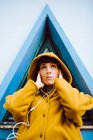 Junge Frau in gelbem warmen Mantel, die Musik hört und in die Kamera schaut, während sie vor einem Dreiecksfenster und einer grauen Hauswand steht — Stockfoto