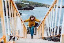 Молодая женщина в жёлтой куртке стоит на ржавой лестнице у залива с размахивающей морской водой — стоковое фото