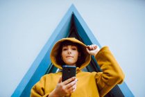 Жінка регулює витяжку жовтого теплого пальто і дивиться на камеру під час перегляду смартфона біля будівлі з трикутним вікном — стокове фото