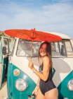 Tanned woman holding red umbrella and smiling near car at sandy beach in bright day — Stock Photo