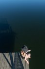 Вид зверху на невпізнавану вдумливу жінку в чорному купальнику та капелюсі, що сидить на дерев'яному пірсі та захоплюючий вид на озеро на прозорому блакитному небі та лісовому тлі — стокове фото