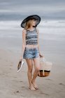 Привлекательная женщина в черной шляпе, держащая пляжную сумку и обувь, наслаждаясь живописным видом на океан — стоковое фото