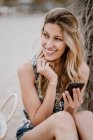 Jovem fêmea atraente com cabelos longos relaxando na praia e usando telefone celular no dia de verão, enquanto olha para longe — Fotografia de Stock