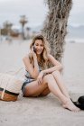 Joven atractiva hembra con el pelo largo relajándose en la orilla del mar y hablando por teléfono móvil en el día de verano - foto de stock