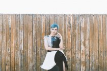 Jeune femme aux cheveux bleus courts portant une robe futuriste et regardant la caméra tout en se tenant près d'une clôture en bois minable — Photo de stock