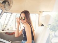 Vue latérale de la jeune femme souriante utilisant des lunettes de soleil assis devant dans la voiture en regardant la caméra — Photo de stock