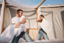 Joyeux jeune couple s'amuser pendant la bataille d'oreillers sur le lit dans une grande tente avec toit transparent — Photo de stock