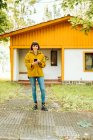 Mujer joven en traje casual sonriendo y navegando teléfono inteligente, mientras que de pie en el camino de baldosas fuera encantadora casa de campo en el día de otoño en el campo - foto de stock