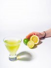 Main féminine tenant le citron coupé en deux et les citrons verts près du verre de limonade jaune boisson sur fond blanc — Photo de stock
