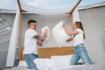 Веселая молодая пара веселится во время боя подушками на кровати в большой палатке с прозрачной крышей — стоковое фото