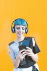Mulher informal alegre em fones de ouvido navegando smartphone e ouvir música enquanto está de pé contra a parede amarela vívida — Fotografia de Stock