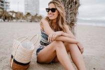 Mujer rubia en gafas de sol negras sentada en la arena con bolsa de verano y mirando hacia otro lado - foto de stock