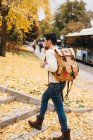 Hombre joven guapo con peinado de moda caminando sobre hojas de otoño y hablando en el teléfono inteligente en el día - foto de stock