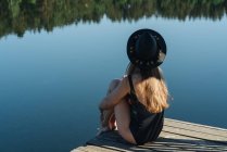 Rückansicht einer unkenntlich gemachten, nachdenklichen Frau in schwarzem Badeanzug und Hut, die auf einem hölzernen Steg sitzt und den Blick auf den See bei klarem blauem Himmel und Waldhintergrund bewundert — Stockfoto