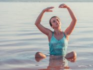 Nachdenkliches Mädchen, das mit erhobenen Händen im Wasser sitzt und aufblickt — Stockfoto