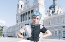 Young woman in trendy futuristic dress holding hand on waist and touching short blue hair while standing on city street — Stock Photo