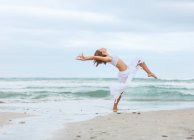 Привлекательная женщина в белом наряде танцует на песке возле моря — стоковое фото