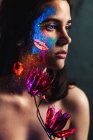 Portrait de belle jeune femme couverte de peinture lumineuse sur le visage tenant une fleur et regardant loin — Photo de stock