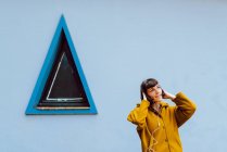 Young woman in yellow warm coat smiling and listening to the music and looking away while standing against triangle window — Stock Photo