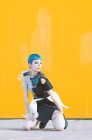 Jeune femme en robe futuriste tendance regardant vers le bas tout en s'asseyant sur les genoux sur le trottoir contre un mur jaune vif — Photo de stock