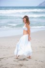 Усміхнена молода жінка в білому вбранні танці на піску біля розмахуючи морем — стокове фото