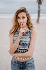 Fröhliche blonde Frau in buntem Oberteil und Jeanshosen macht Schweigegegeste mit der Hand am Strand — Stockfoto