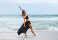Привлекательная женщина в черном наряде танцует на песке у моря — стоковое фото