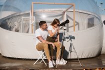 Stylish woman sitting on boyfriend lap and looking through telescope at sky near bubble hotel — Stock Photo