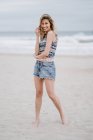 Fröhliche blonde Frau in buntem Top und Jeanshosen lächelnd und in die Kamera blickend, während sie sich am Strand entspannt — Stockfoto