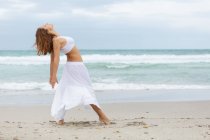 Elegant woman in white outfit dancing on sand near waving sea — Stock Photo