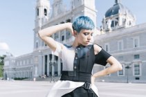 Молодая женщина в модном футуристическом платье, держась за талию и прикасаясь к коротким голубым волосам стоя на городской улице — стоковое фото