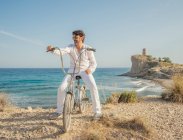 Maturo uomo barbuto sportivo utilizzando occhiali da sole mentre in bicicletta sul mare con erba secca su sfondo di incredibile paesaggio marino turchese in giornata luminosa — Foto stock