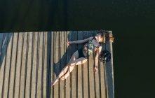 Vue du dessus de la belle femme en maillot de bain noir et chapeau couché sur la jetée en bois du lac sur ciel bleu clair et fond de forêt — Photo de stock